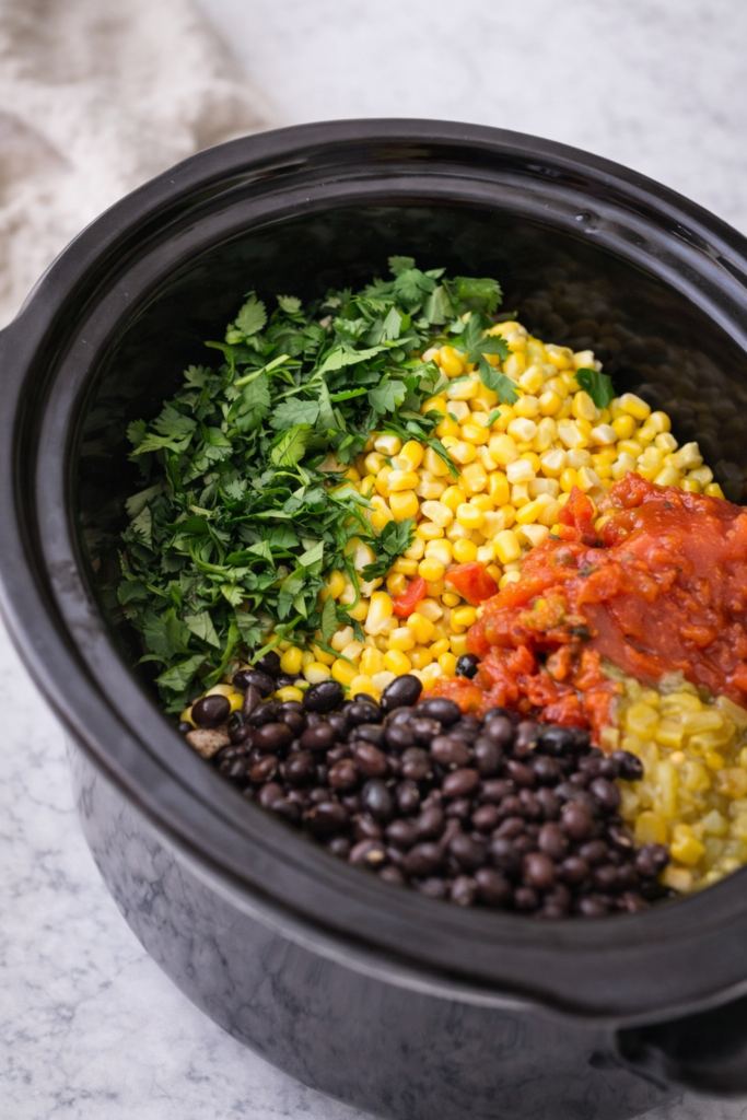 Ingredients for crockpot chicken tortilla soup layered in a slow cooker including black beans, corn, diced tomatoes, and fresh cilantro.