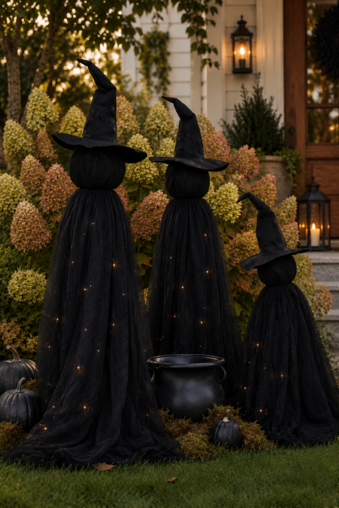 Three DIY light-up Halloween witches made with black gauze fabric and fairy lights displayed in front of blooming Limelight hydrangeas in soft pink and green fall colors with a black cauldron and pumpkins.