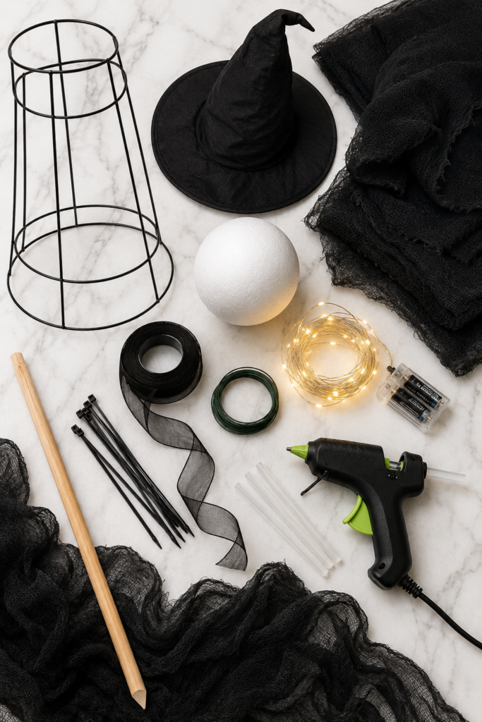 Flat lay of supplies needed to make DIY light-up Halloween witches, including a tomato cage, black gauze fabric, witch hat, foam ball, fairy lights, hot glue gun, floral wire, ribbon, zip ties, and wooden dowel on a white marble countertop.