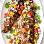 Juicy grilled mojo pork tenderloin topped with fresh mango salsa on a white platter over a marble countertop.