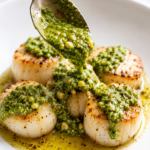 Spoon pouring homemade walnut basil pesto over golden pan seared scallops on a white plate with a buttery sauce and fresh basil flavors.