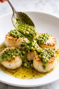 Spoon pouring homemade walnut basil pesto over golden pan seared scallops on a white plate with a buttery sauce and fresh basil flavors.