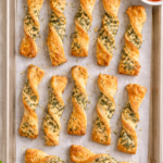Overhead flat lay of baked herb cheese twists on a parchment-lined baking sheet, golden brown and topped with Parmesan and dried herbs.