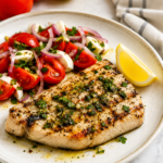 Sicilian style grilled swordfish topped with garlic herb olive oil and served with fresh tomato and red onion salad on a white plate.