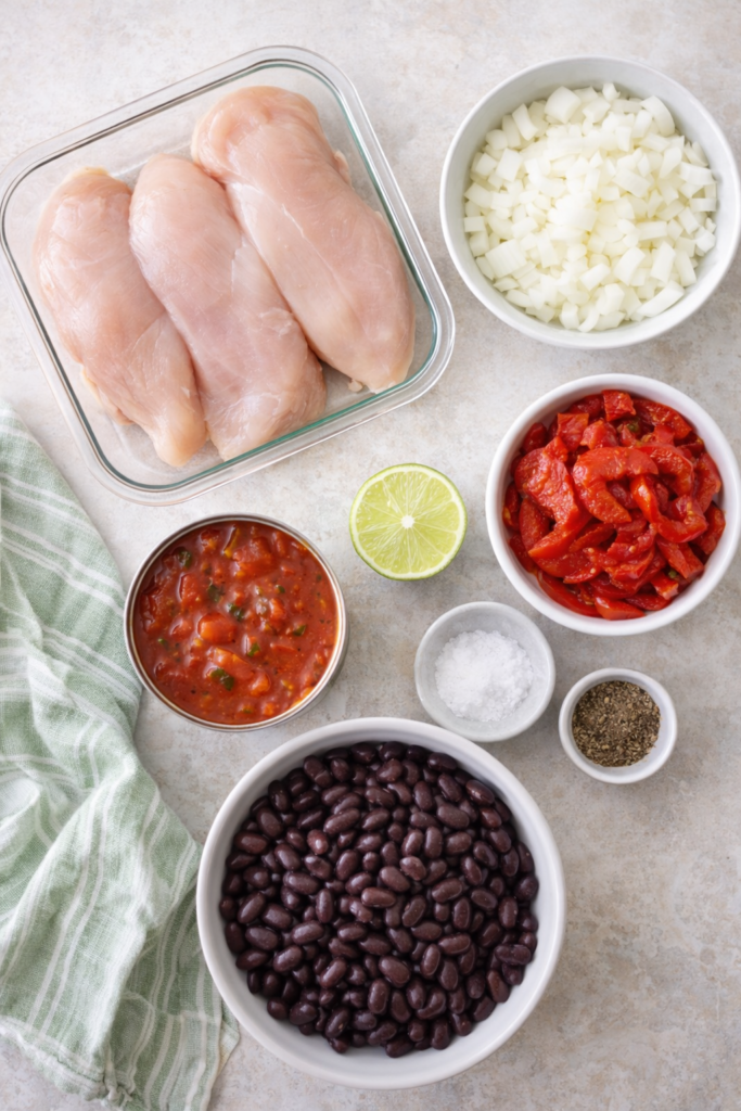 Flat lay of ingredients for Southwestern chicken with black beans including raw chicken breasts, black beans, roasted red peppers, tomatoes, onion, lime, salt, and pepper