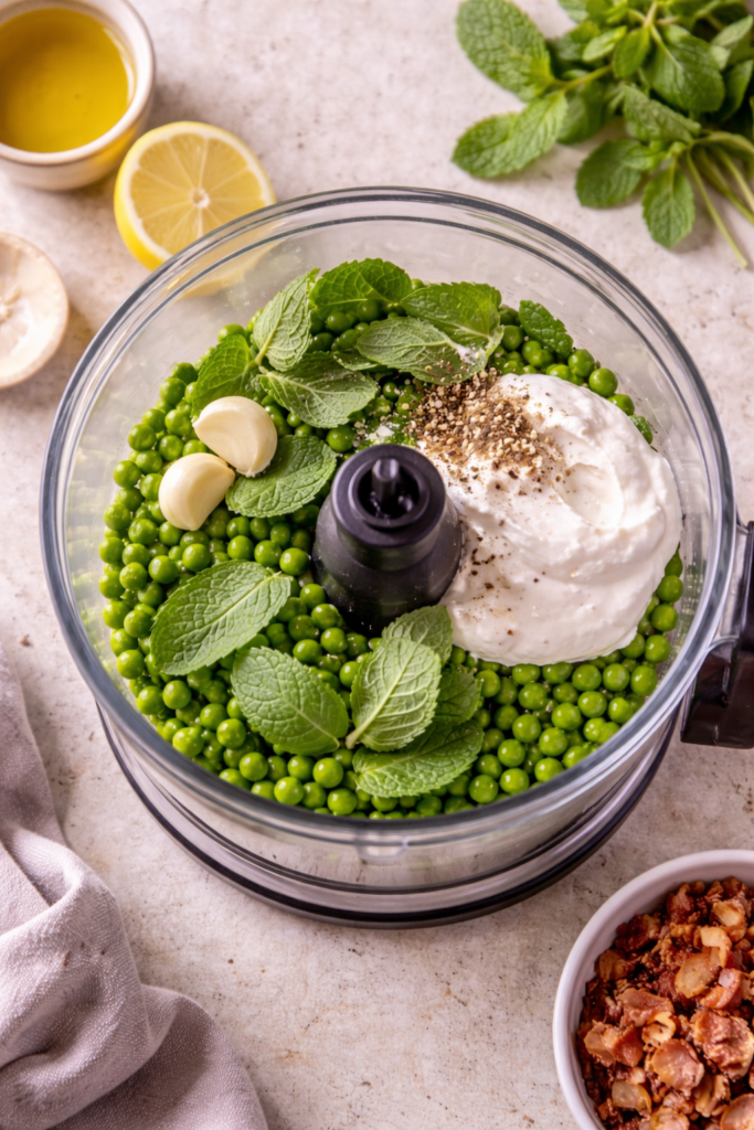 Ingredients for creamed pea and mint sauce in a food processor before blending, including fresh peas, mint leaves, garlic, sour cream, salt, and pepper.