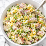 Creamy summer potato salad with red potatoes, eggs, peas, celery, carrots, and a tangy Dijon dressing served in a white bowl.