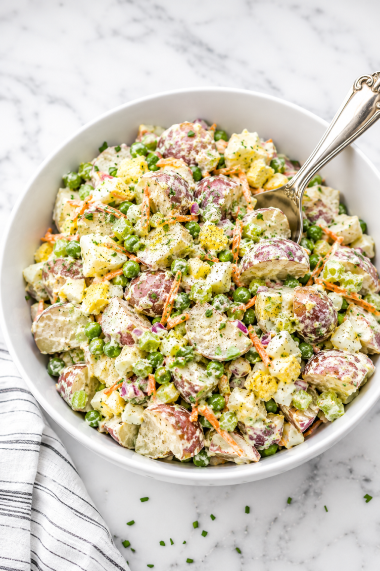 Creamy summer potato salad with red potatoes, eggs, peas, celery, carrots, and a tangy Dijon dressing served in a white bowl.