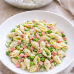 Creamy ham and pea pasta salad in a white bowl with shell pasta, green peas, diced ham, and poppy seeds, styled in a bright bird’s-eye view with a serving bowl blurred in the background.