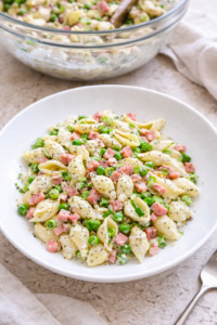 Creamy ham and pea pasta salad in a white bowl with shell pasta, green peas, diced ham, and poppy seeds, styled in a bright bird’s-eye view with a serving bowl blurred in the background.