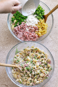 Bird’s-eye view of creamy ham and pea pasta salad being mixed in a glass bowl with shell pasta, diced ham, green peas, scallions, and creamy dressing being poured in.