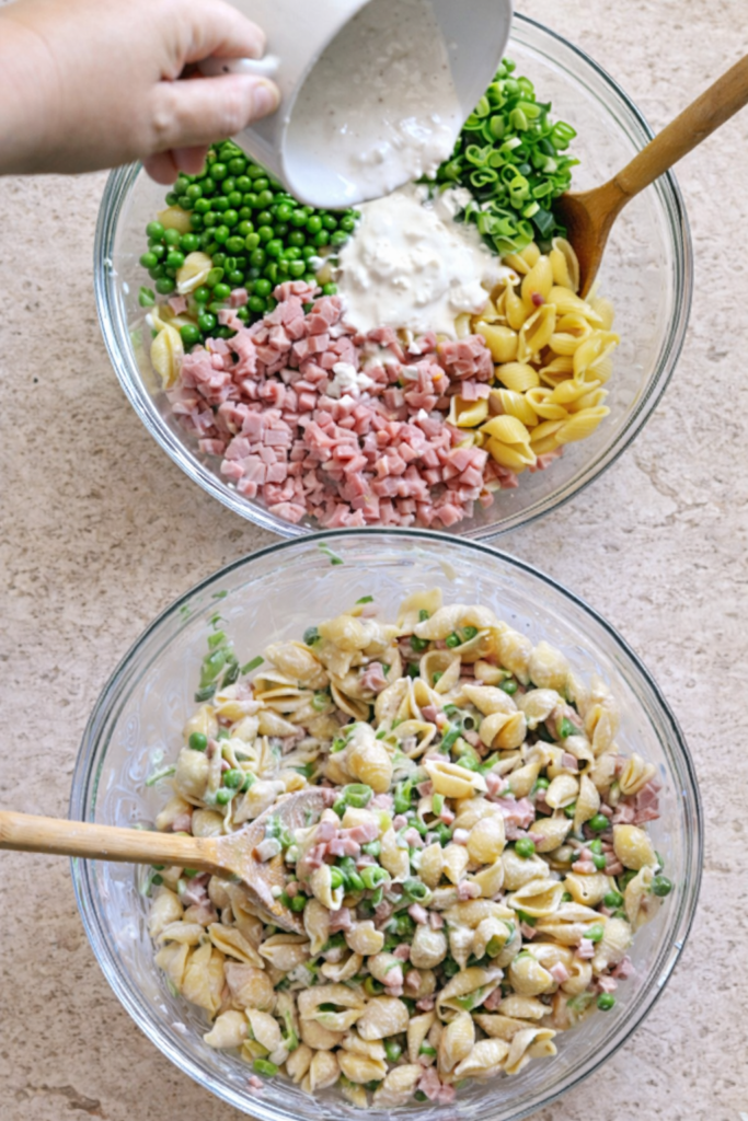 Bird’s-eye view of creamy ham and pea pasta salad being mixed in a glass bowl with shell pasta, diced ham, green peas, scallions, and creamy dressing being poured in.