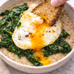 Toast dipping into a runny egg yolk over savory oatmeal with spinach in a rustic pottery bowl