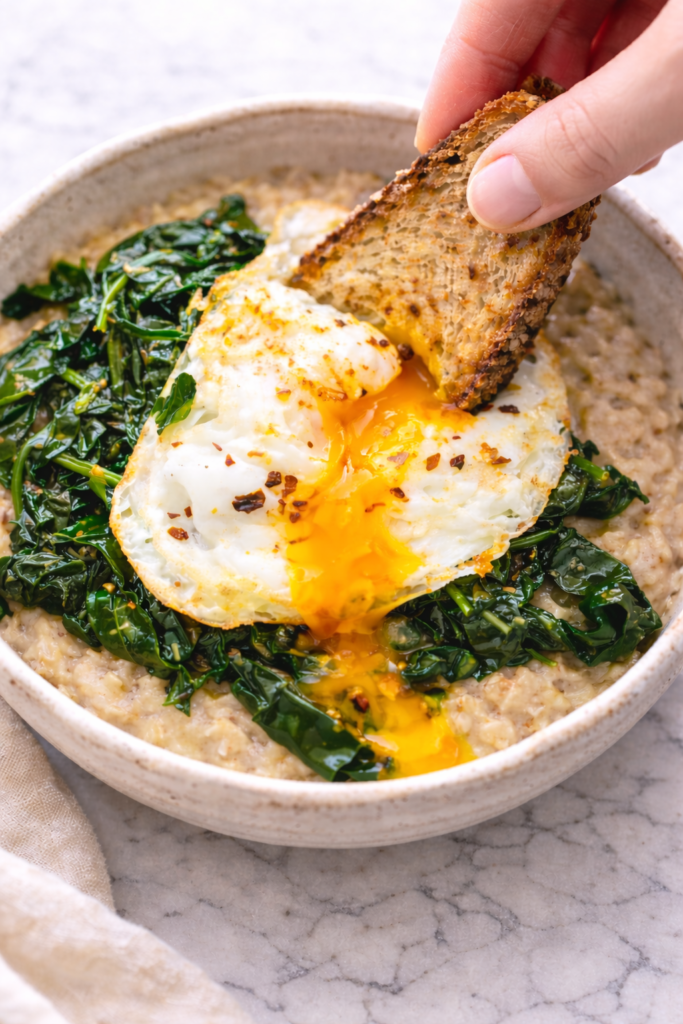 Toast dipping into a runny egg yolk over savory oatmeal with spinach in a rustic pottery bowl