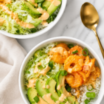 Bang Bang shrimp bowls with crispy shrimp, avocado, lettuce, rice, and spicy sauce served in white bowls on a marble countertop.