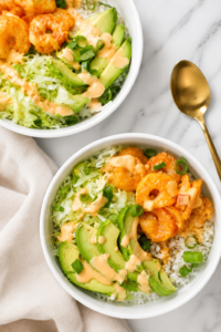 Bang Bang shrimp bowls with crispy shrimp, avocado, lettuce, rice, and spicy sauce served in white bowls on a marble countertop.