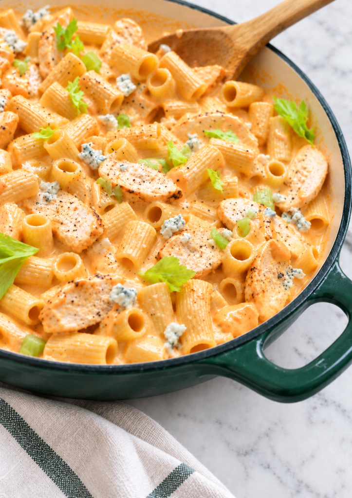 Close side view of creamy garlic Buffalo chicken pasta with rigatoni, chicken, celery, and blue cheese in a dark green enamel skillet.