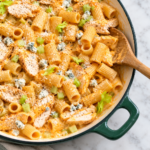 Creamy garlic Buffalo chicken pasta with rigatoni, tender chicken, celery, blue cheese crumbles, and Parmesan in a dark green enamel skillet.