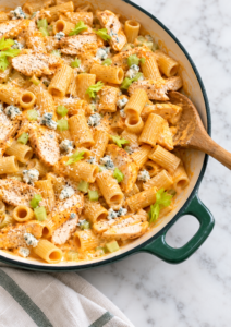 Creamy garlic Buffalo chicken pasta with rigatoni, tender chicken, celery, blue cheese crumbles, and Parmesan in a dark green enamel skillet.