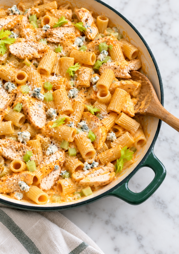Creamy garlic Buffalo chicken pasta with rigatoni, tender chicken, celery, blue cheese crumbles, and Parmesan in a dark green enamel skillet.
