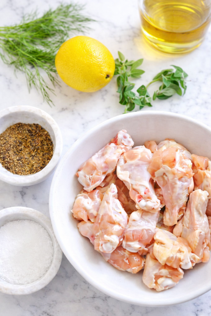 Raw chicken wings in a white bowl with lemon, fresh herbs, olive oil, salt, and pepper