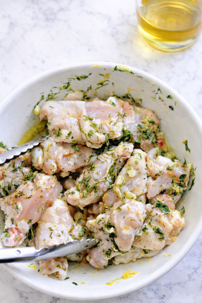 Raw chicken wings coated in lemon herb marinade in a white bowl with tongs