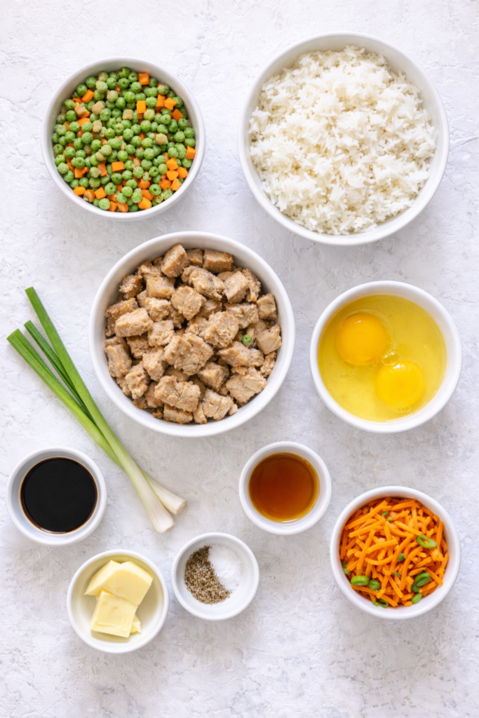 Flat lay of ingredients for pork fried rice including cooked rice, diced pork, peas and carrots, eggs, soy sauce, sesame oil, butter, and green onions on a marble surface.