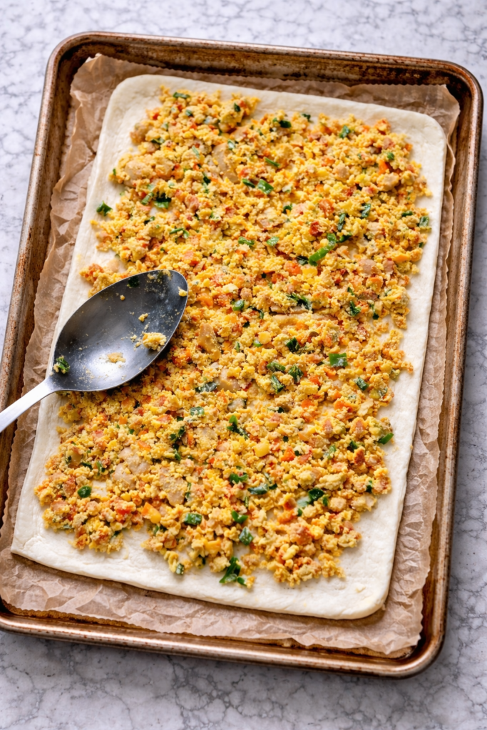 Breakfast burrito bun filling spread over dough on a parchment-lined baking sheet