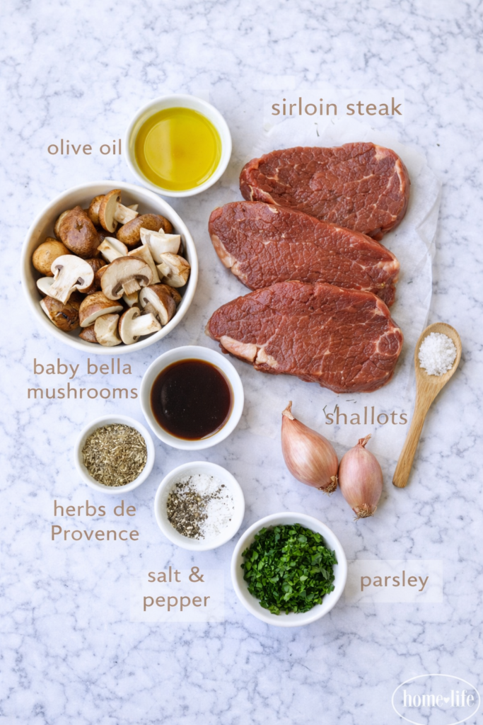 Flat lay of ingredients for pan-seared sirloin steak including raw sirloin, baby bella mushrooms, shallots, olive oil, herbs de Provence, salt, pepper, and fresh parsley on a marble surface.