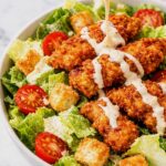 Spicy chicken Caesar salad with crispy fried chicken, romaine lettuce, tomatoes, croutons, and Caesar dressing being poured over the top