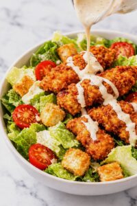 Spicy chicken Caesar salad with crispy fried chicken, romaine lettuce, tomatoes, croutons, and Caesar dressing being poured over the top