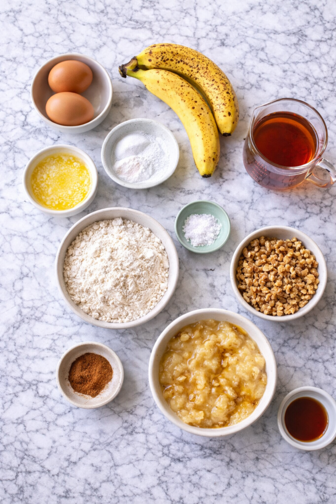 Flat lay of maple banana muffin ingredients including ripe bananas, maple syrup, flour, eggs, butter, and cinnamon on a marble countertop