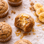 Maple banana muffins on crinkled parchment paper with butter and maple syrup dripping and sliced bananas on the side