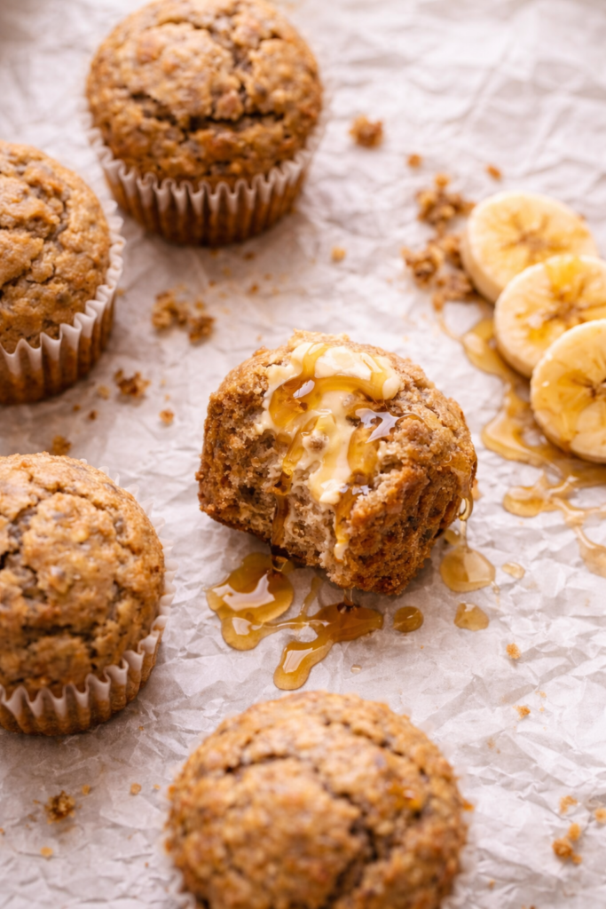 Maple banana muffins on crinkled parchment paper with butter and maple syrup dripping and sliced bananas on the side