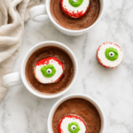 Three white mugs of hot chocolate topped with homemade marshmallow eyeballs on a marble counter.