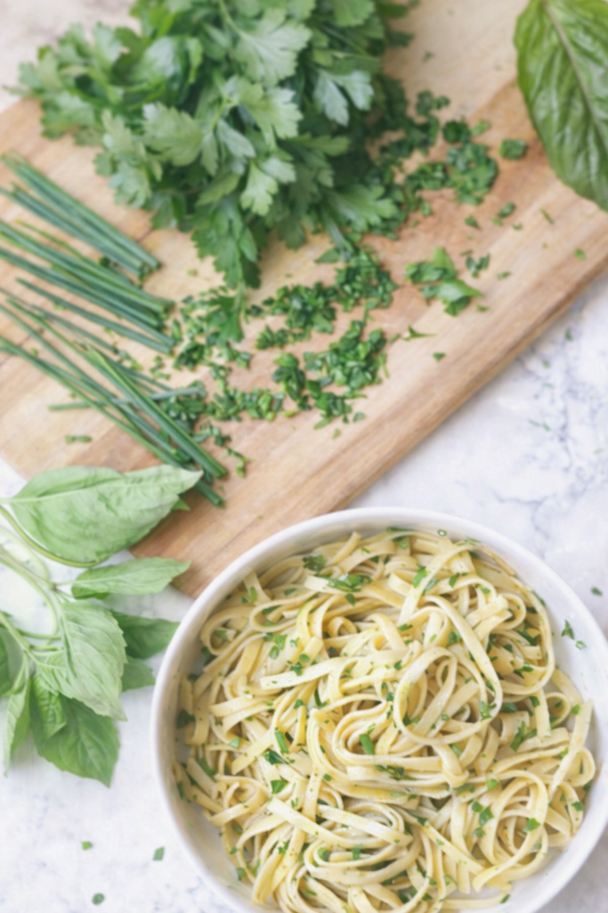 Overhead view of fresh herb fettuccine Alfredo with parsley, chives, and basil on a cutting board beside a bowl of creamy pasta
