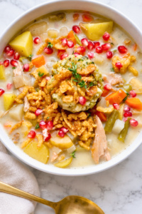 Close-up of creamy turkey chowder with potatoes, carrots, and green beans topped with stuffing, crispy fried onions, pomegranate seeds, and thyme.