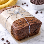 Moist chocolate chip banana bread loaf wrapped in parchment paper with ripe bananas and chocolate chips in the background