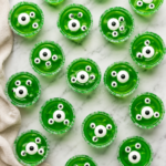 Bird's-eye view of spooky Monster Jello Shots made with bright green gelatin, candy eyeballs, and green sugar rims arranged on a white marble countertop for a Halloween party.