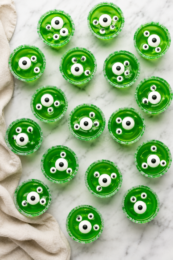 Bird's-eye view of spooky Monster Jello Shots made with bright green gelatin, candy eyeballs, and green sugar rims arranged on a white marble countertop for a Halloween party.