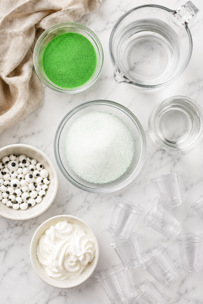 Flat lay of ingredients for Monster Jello Shots including lime gelatin, vodka, water, candy eyeballs, green sanding sugar, whipped cream, and plastic shot cups on a white marble countertop.