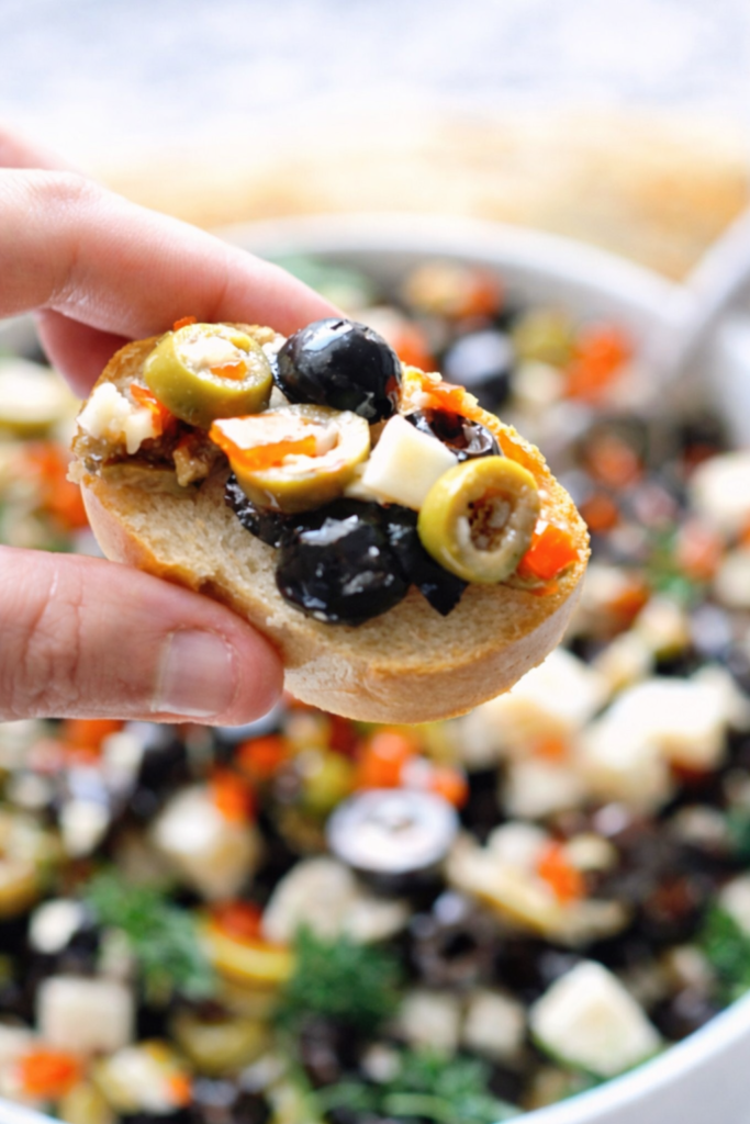 close up of olive and parmesan salad dip served on toasted baguette slice with black and green olives, cheese cubes, and herbs over a bowl on a marble counter