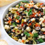 chunky olive parmesan salad dip with black and green olives, garlic, herbs, and cheese cubes served in a white bowl on a marble counter with bread slices
