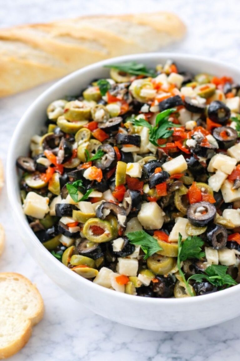 chunky olive parmesan salad dip with black and green olives, garlic, herbs, and cheese cubes served in a white bowl on a marble counter with bread slices