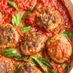 Spicy Italian meatballs in marinara sauce with fresh basil and red pepper flakes in a silver skillet on a white marble countertop.