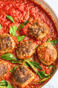 Spicy Italian meatballs in marinara sauce with fresh basil and red pepper flakes in a silver skillet on a white marble countertop.