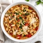 Homemade Italian chicken noodle soup in a white bowl topped with Parmesan and fresh parsley on a white marble countertop with a neutral kitchen towel.