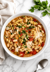 Homemade Italian chicken noodle soup in a white bowl topped with Parmesan and fresh parsley on a white marble countertop with a neutral kitchen towel.
