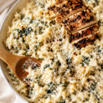Creamy spinach artichoke chicken pasta in a white skillet topped with sliced blackened chicken on a marble countertop with a wooden spoon and neutral linen cloth.