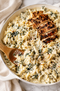 Creamy spinach artichoke chicken pasta in a white skillet topped with sliced blackened chicken on a marble countertop with a wooden spoon and neutral linen cloth.